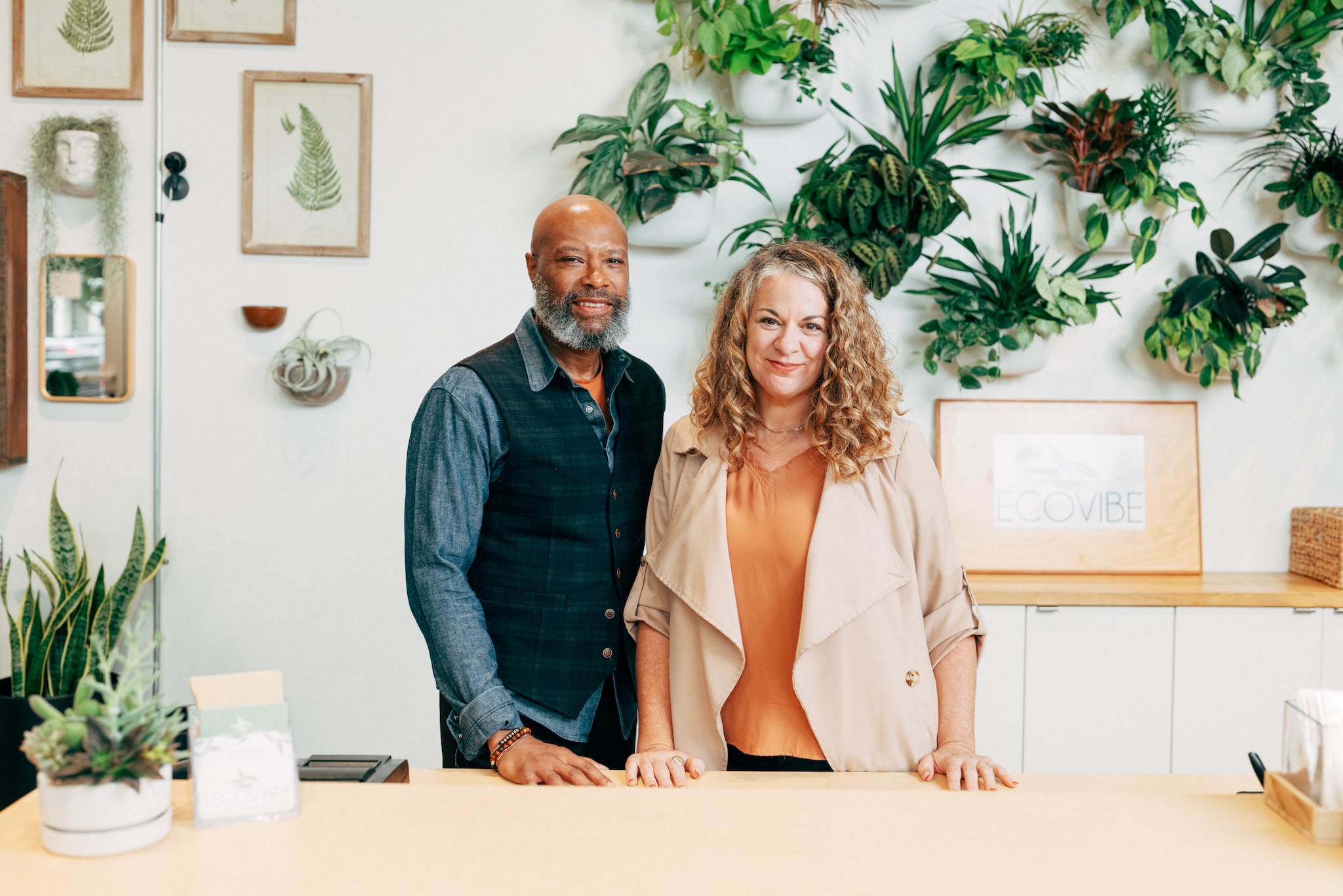 two shopkeepers stand behind a counter with plants in the background