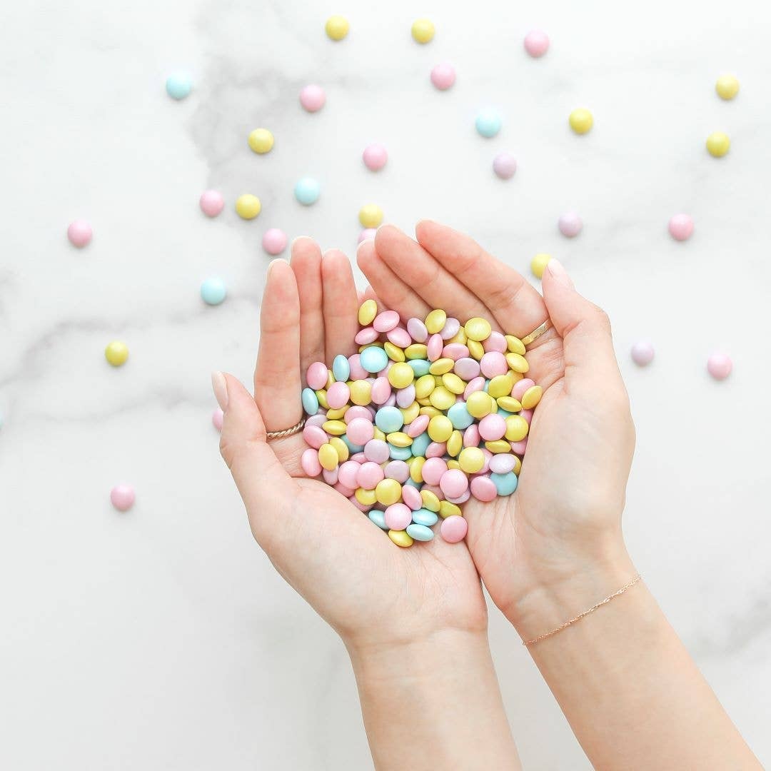 hands cup pastel colored candy coated chocolates in front of a marble surface